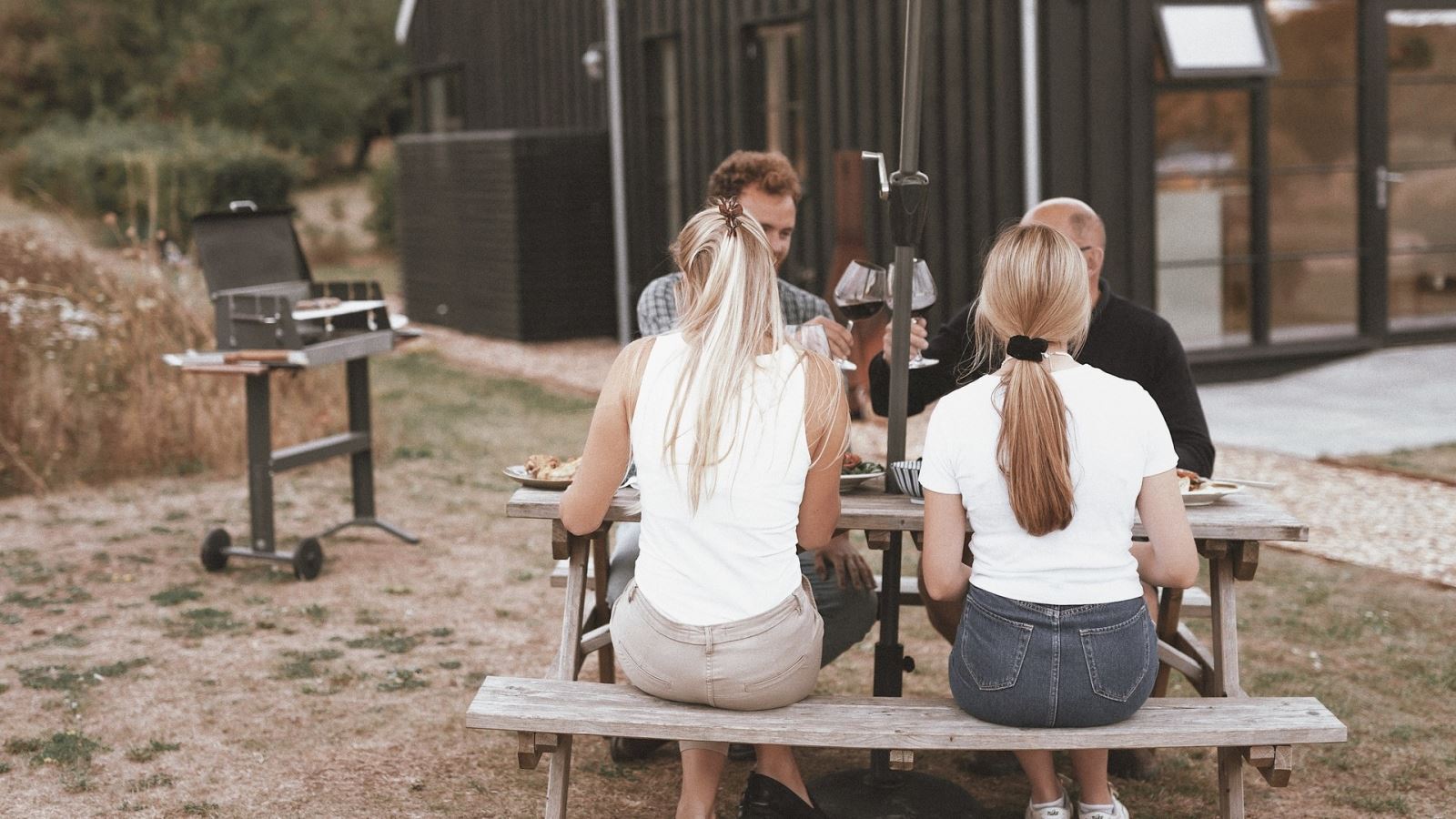 Four people sat outside on a picnic table drinking wine in front of a barn house