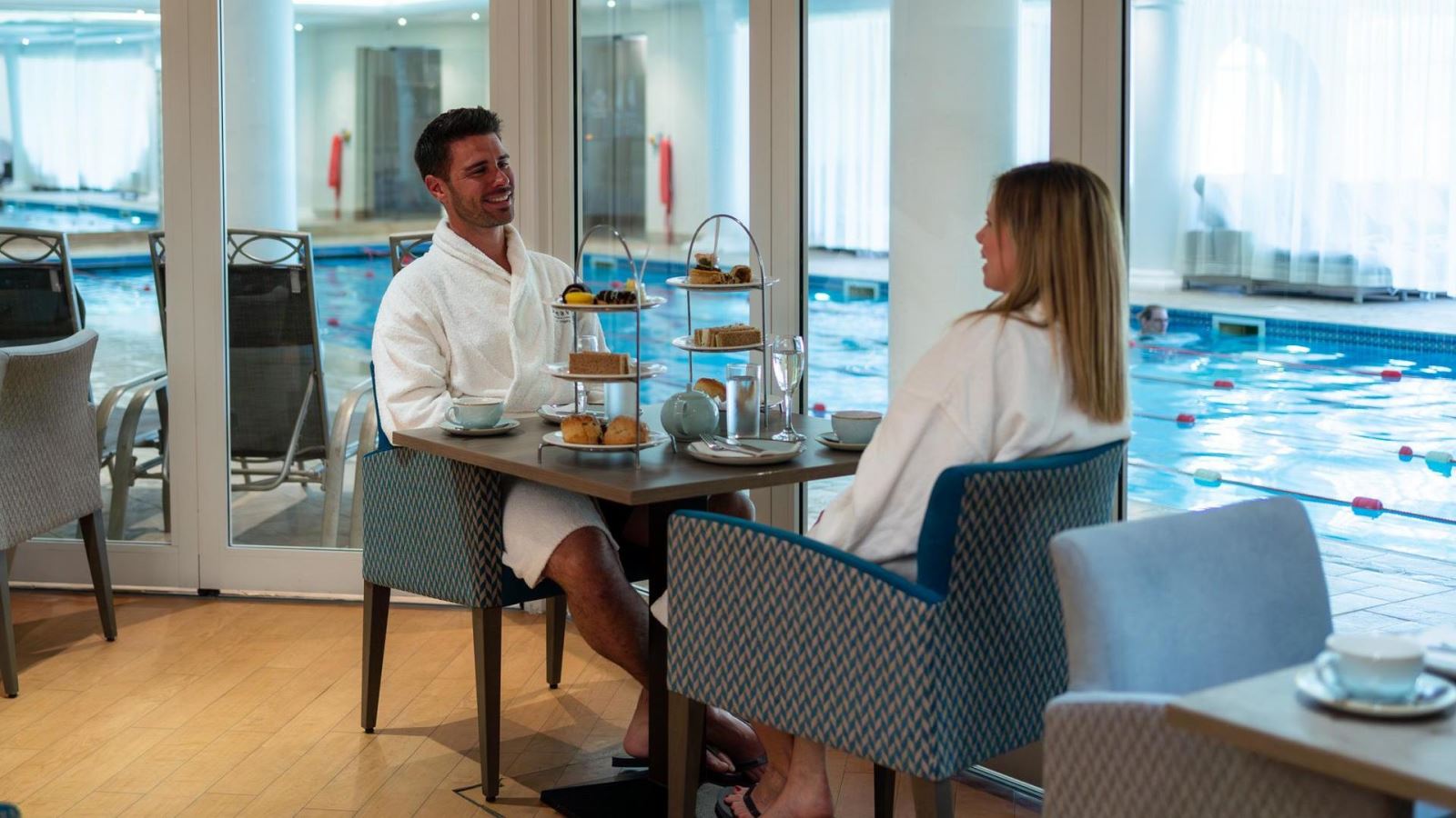 Man and woman having afternoon tea at Stoke by Nayland spa wearing white robes with a swimming pool view